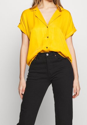 Chemise jaune à manches courtes avec col, boutonnée et coupe ample, associée à un jean noir taille haute. Texture lisse, détails minimalistes.