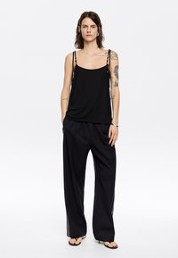 Top nero senza maniche con spalline regolabili, abbinato a pantaloni neri larghi con strisce bianche ai lati. Sandali piatti completano il look.