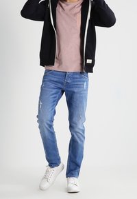 Hombre con jeans rasgados de color azul desgastado, zapatillas blancas, una camiseta rosa y una sudadera con cremallera negra sobre un fondo liso.