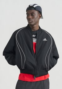 Νέος άνδρας που φοράει μαύρο μπουφάν Adidas, κόκκινο πουκάμισο και μαύρο-άσπρο καπέλο που φοράει ανάποδα, στέκεται με τα χέρια στις τσέπες.