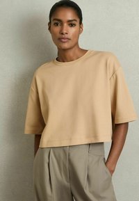 Beige lyhyt crop-top, pehmeää kangasta, pyöreällä kaula-aukolla ja lyhyillä hihoilla, yhdistettynä korkeavyötäröisiin, vaaleanharmaisiin muodikkaisiin housuihin.