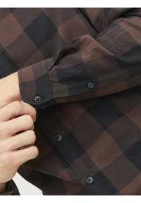 Personne qui ferme le bouton de la manche d'une chemise à carreaux marron et noir à manches longues.