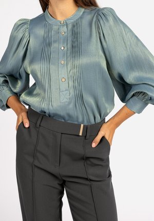 Femme portant un chemisier en satin bleu clair à manches bouffantes et boutons décoratifs, associé à un pantalon noir taille haute avec une ceinture.