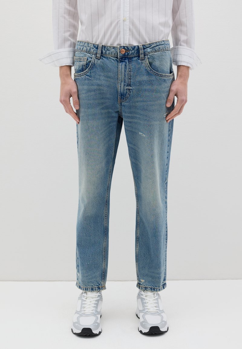 Jean en denim bleu clair avec une coupe droite, des détails déchirés et une fermeture à bouton à la taille. Porté avec des baskets blanches.