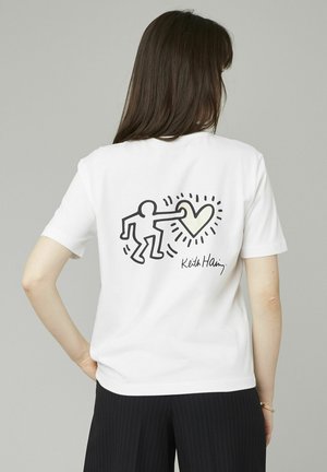 T-shirt en coton blanc avec un dessin en contour noir d'une figure tendant la main vers un cœur jaune, avec "Keith Haring" imprimé en dessous.