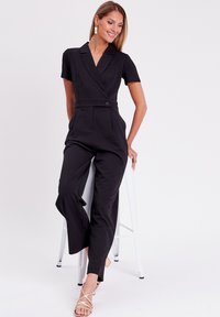 Jumpsuit noir à manches courtes avec un design enveloppé, doté d'un col à revers, d'une taille ceinturée et d'une coupe évasée. Texture lisse et détails minimaux.