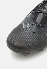 Svart fotbollssko med texturerad syntetisk ovandel med geometriskt mönster, rund tå och svarta snören. Sulan har synliga dobbar.