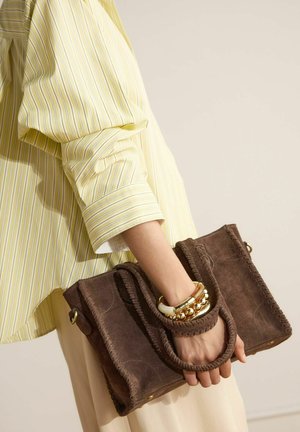 Hand holding a brown suede handbag with braided handles, wearing a yellow striped shirt and gold and white chunky bracelets.