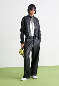 Zwarte leren bomberjack met zilverkleurig jumpsuit, brede zwarte broek en witte sneakers. Houdt een kleine zwart-gele tas met metalen accenten vast.