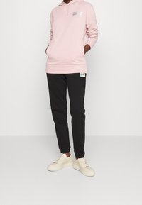 Jasnoróżowa bluza z kapturem z logo na piersiach, zestawiona z czarnymi spodniami dresowymi z metką logo. Model nosi kremowe sneakersy.