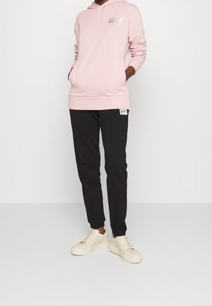Felpa con cappuccio rosa chiaro con logo sul petto, abbinata a pantaloni jogger neri con etichetta del logo. Il modello indossa sneaker color crema.