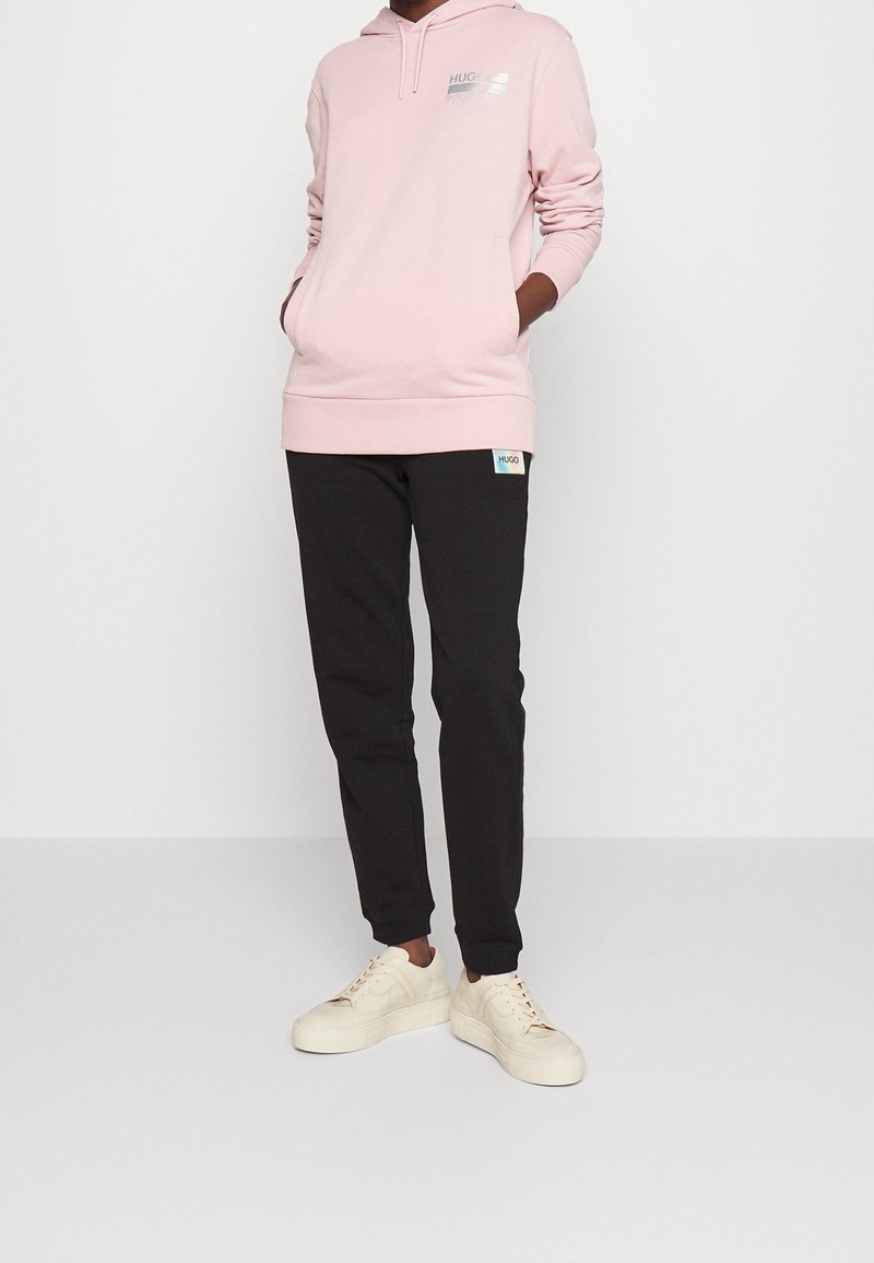 Jasnoróżowa bluza z kapturem z logo na piersiach, zestawiona z czarnymi spodniami dresowymi z metką logo. Model nosi kremowe sneakersy.