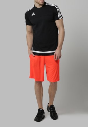 Chemise de sport noire avec des rayures blanches, associée à un short orange vif et des chaussures de sport noires. La tenue présente une coupe ajustée et un tissu respirant.