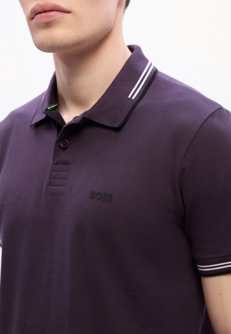 Homme portant un polo violet foncé avec un col et des bordures de manches rayés noir et blanc, arborant un logo "BOSS" noir sur la poitrine.