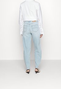 Person som bär ljusblå högmidjade jeans, vit långärmad blus med broderade detaljer, och beige klackar med svarta remmar, står med ryggen mot oss.