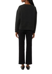 Frau von hinten, die ein schwarzes, strukturiertes Langarmoberteil, gerade schwarze Hosen und schwarze flache Schuhe mit Knöchelriemen trägt.