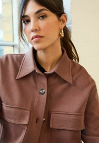 Braune Button-Up-Jacke aus weichem Material mit Kragen, zwei Fronttaschen und Knopfverschluss. Dezente Textur und elegantes Design.
