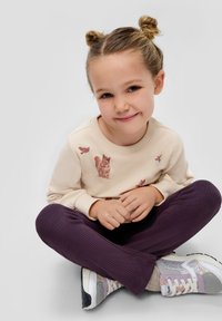 Lichtbeige sweatshirt met dierenprints, paarse geribde leggings en grijze sneakers met sprankelende accenten. Kind zit met handen op de knieën.