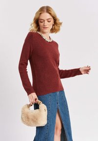 Bordo džemperis izgatavots no mīksta materiāla, ar garām piedurknēm un ribotām aprocēm, kombinēts ar džinsu svārkiem ar priekšējo šķirbu, un tur rokās beigu mākslīgās kažokādas somiņu.