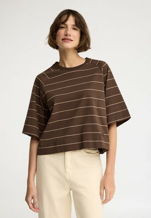 Młoda kobieta o krótkich brązowych włosach, ubrana w luźną bluzkę w brązowe paski z krótkim rękawem oraz jasnobeżowe spodnie, lekko się uśmiecha.