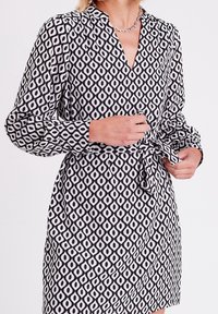 Robe à motifs noir et blanc avec manches longues, col et taille nouée. Présente un motif géométrique en diamant et une texture de tissu lisse.