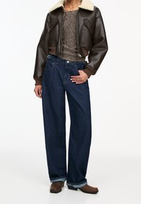 Bruine leren cropped jas met schapenvachtkraag, grijze gebreide trui, losse donkerblauwe jeans en bruine cowboylaarzen met een lage hak.