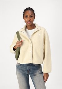Kermainen fleece-takki, joka on lyhennetty muotoilultaan, napsautettavia nappeja ja korkea kaulus. Yhdistetty sinisiin farkkuihin ja vihreään olkalaukkuun.