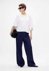 Biała bluzka z krótkim rękawem, szerokie spodnie w niebieskie paski, czarna torebka i czarne okulary przeciwsłoneczne. Teksturowana tkanina o luźnym kroju.