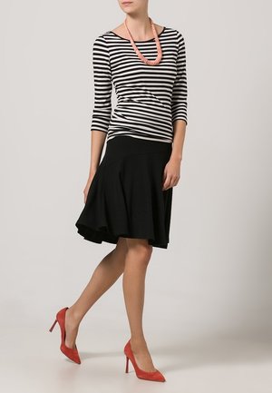 Schwarzer ausgestellter Rock kombiniert mit einem schwarz-weiß gestreiften Oberteil. Das Model trägt korallenrote High Heels und eine passend korallenrote Halskette.