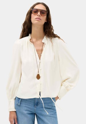 Frau mit übergroßen Schildpatt-Sonnenbrillen, einer weißen Bluse mit langen Ärmeln und Knöpfen und blauen Jeans, die mit einer Hand in der Tasche posiert.