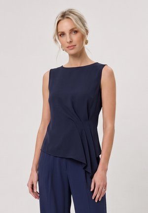 Donna bionda che indossa un top blu navy senza maniche con drappeggio asimmetrico e pantaloni blu navy abbinati, in piedi su sfondo chiaro.