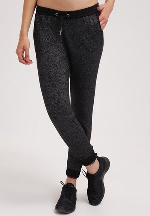 Osoba ubrana w ciemnoszare, teksturowane spodnie dresowe z czarnym elastycznym pasem i ściągaczami, czarne buty sportowe, stojąca ze skrzyżowanymi nogami.