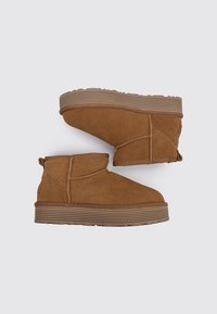 Botas de tobillo de ante marrón con una suela de goma texturizada, que presentan un diseño sencillo y detalles de costura en las costuras laterales, dispuestas en ángulo para su exhibición.