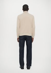 Beige långärmad topp med hög krage, parat med mörkblå raka jeans och svarta skor, sedd bakifrån.