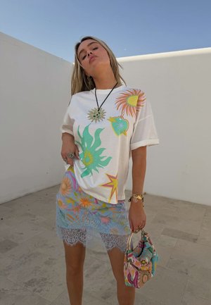 Kobieta w białej koszuli z kolorowymi nadrukami słońca i gwiazd, przezroczystą niebieską spódnicę z koronkowym obszyciem, trzymająca na zewnątrz wielokolorową torebkę w wzory.