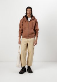 Brown zip-up hoodie with front pockets, beige loose-fitting cargo pants, and black lace-up boots. Holds a light beige tote bag. Simple background.