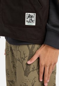 Schwarze Canvasjacke mit einem weißen Aufnäher, der ein Bigfoot-Motiv zeigt. Olivgrüne Hose mit einem dunklen Baumzweigmuster.