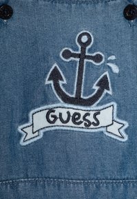 Il tessuto di denim presenta un'ancora ricamata di colore navy con un nastro su cui è scritto "Guess." Lo sfondo è un denim leggero azzurro e testurizzato.