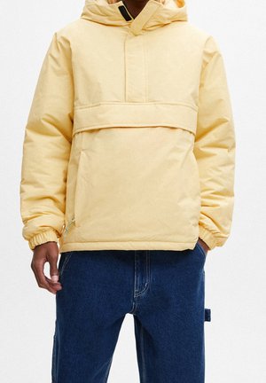Mann trägt eine hellgelbe Kapuzen-Steppjacke mit elastischen Bündchen und dunkelblaue Jeans mit einer Seitenschlaufe.