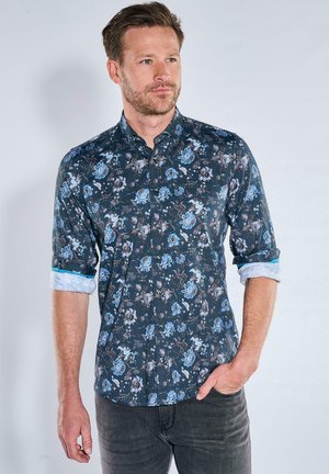 Mann mit hochgekrempelten Ärmeln, der ein dunkelblaues, geblümtes Hemd mit Knöpfen und dunkle Jeans trägt, blickt leicht seitlich mit der Hand in der Tasche.