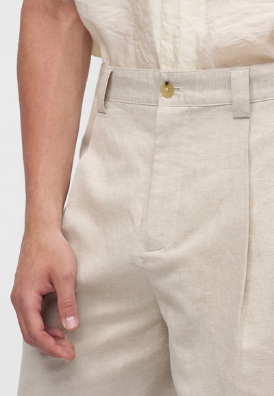 Gros plan d'une personne portant un pantalon en lin beige avec un bouton et des passants pour ceinture, et une chemise beige clair partiellement rentrée.
