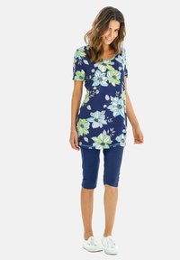 Bloemige tuniektop met blauwe en groene bloemen op een marineblauwe achtergrond, gecombineerd met marineblauwe knielange Capri-broeken en witte sneakers.