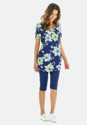 Bloemige tuniektop met blauwe en groene bloemen op een marineblauwe achtergrond, gecombineerd met marineblauwe knielange Capri-broeken en witte sneakers.