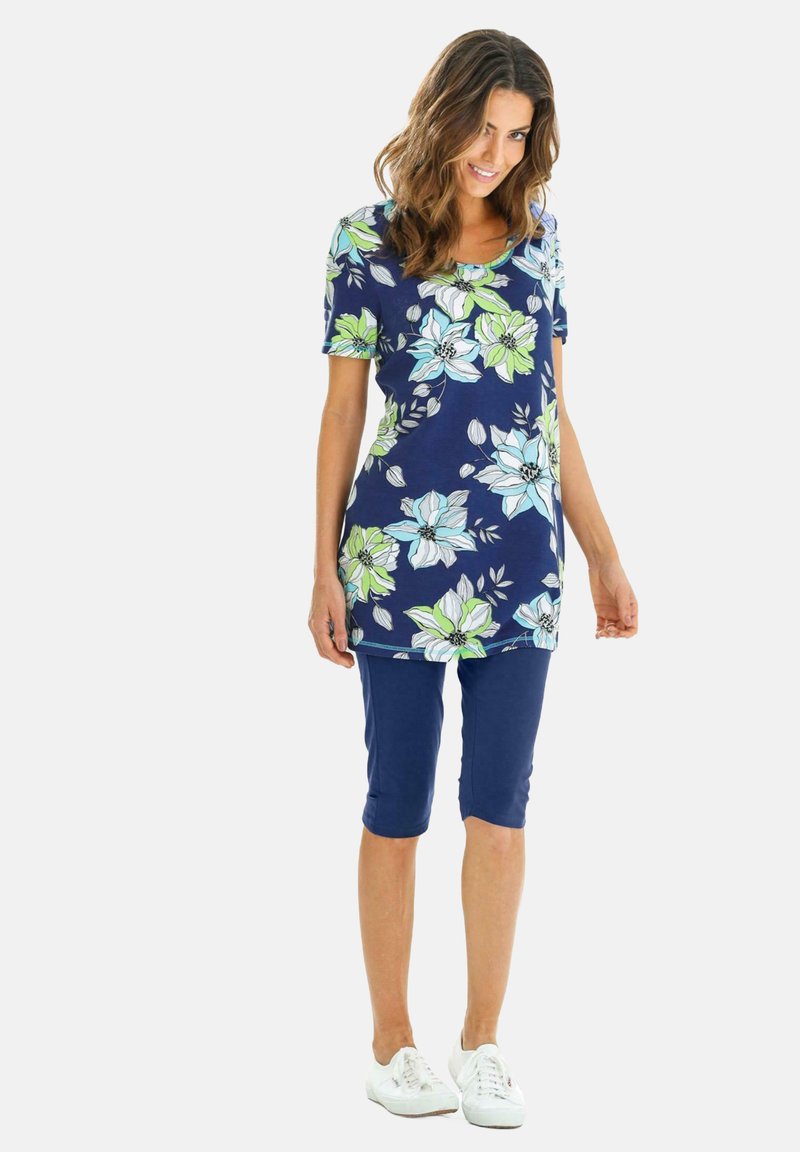 Bloemige tuniektop met blauwe en groene bloemen op een marineblauwe achtergrond, gecombineerd met marineblauwe knielange Capri-broeken en witte sneakers.