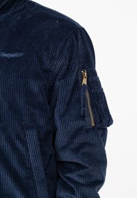 Marineblauwe corduroy jas met een ritszak op de linker mouw, voorzien van een geribbelde textuur en subtiele geborduurde details op de borst.