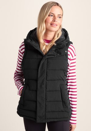 Woman wearing a black quilted sleeveless vest over a long-sleeve pink and white striped shirt, hands in vest pockets, smiling slightly.