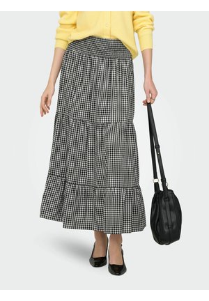 Femme portant une jupe à volants à carreaux noirs et blancs, un pull jaune, des chaussures noires à bout pointu, tenant un sac à main en cuir noir par la anse.