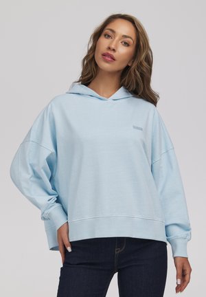 Kobieta z długimi falowanymi włosami, ubrana w jasnoniebieską, oversize'ową bluzę z kapturem i ciemne dżinsy, stojąca na jednolitym neutralnym tle.