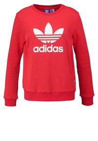 Röd bomullströja med långa ärmar, ribbade muddar och nederkant. Har en stor vit Adidas-logotyp och klassisk tre-randig design på bröstet.