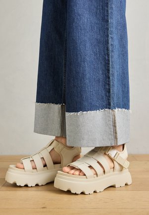 Beige plattform sandaler med tykke såler har en stropdesign og justerbare ankelstropper. Båret med brede denimbukser, har den ene kanten slitt.